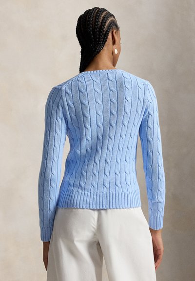 Žena s vrkočmi, oblečená v svetlomodrom sveteri s vrkočovým vzorom a bielych nohaviciach, zobrazená zozadu na neutrálnom pozadí.