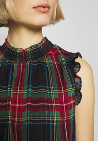 Haut sans manches en tartan avec un col smocké, des détails volantés sur les épaules et une palette de couleurs rouge, vert, jaune et noir.
