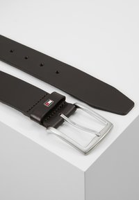Cinturón de cuero marrón con hebilla de metal plateado y diseño rectangular. Cuenta con múltiples agujeros y un acento con la marca cerca de la hebilla.