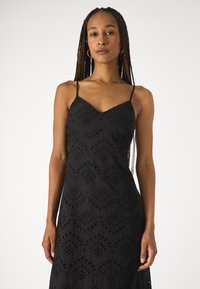 Vestido preto sem mangas com decote em V, apresentando bordados intrincados e padrões recortados por todo o tecido. Textura suave.