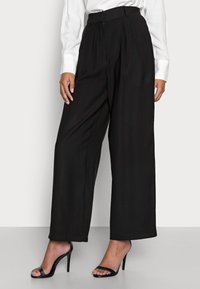 Černé kalhoty s širokými nohavicemi a vysokým pasem, s nařaseným předním dílem a hladkým materiálem, doplněné černými sandály na vysokém podpatku.