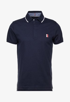 Polo à manches courtes bleu marine avec des bordures blanches au col et aux manches, avec un petit logo rectangulaire rouge, blanc et bleu sur la poitrine.