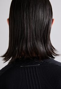 Crna pletena odjeća s teksturiranim uzorcima, s rebrastim detaljima i stražnjim šavom. Glatka, ravna kosa koja pada malo ispod ramena.