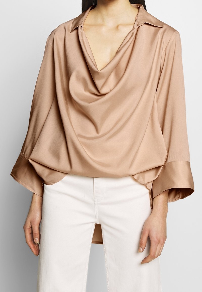 Beige, satiiniblouse, jossa on laskostettu pääntie, taitettavat hihansuut ja rento istuvuus. Ominaisuuksiin kuuluu pitkät hihat ja epäsymmetrinen helma.