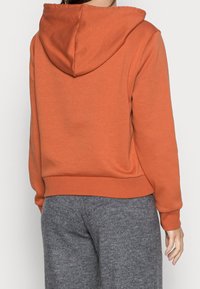 Oranžová mikina s kapucí a stahovací šňůrkou, s žebrovanými manžetami. Tkanina vypadá měkká s uvolněným střihem, kombinována se šedými strukturovanými kalhotami.