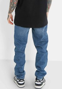 Blå denimjeans med en avslappnad passform och en urtvättad textur. Bärs med svarta och vita Nike-sneakers. Synlig tatuering på armen.