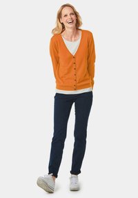 Oranje cardigan met V-hals en knoopsluiting, over een wit shirt, gecombineerd met donkere jeans en witte sneakers.