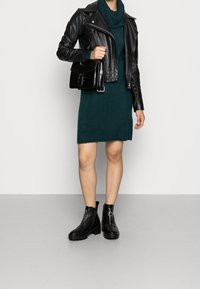 Giubbotto in pelle nera da motociclista sopra un vestito di maglia color turchese. Stivali neri alla caviglia con cerniere e tacchi grossi. Piccola borsa a tracolla nera in vernice con catena.