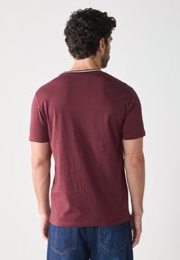 Camiseta de algodón burdeos con cuello redondo y mangas cortas; presenta una franja horizontal blanca alrededor del cuello y un corte clásico.