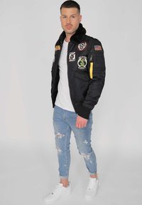 Svart bomberjacka med lappar, dragkedjefickor och pälskrage, parat med slitna ljusblå jeans och vita sneakers.