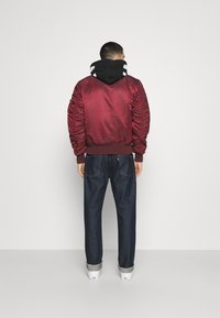 Giacca bomber in satin bordeaux con polsini e orlo a coste, abbinata a jeans di denim scuri arrotolati alla caviglia e sneakers bianche.