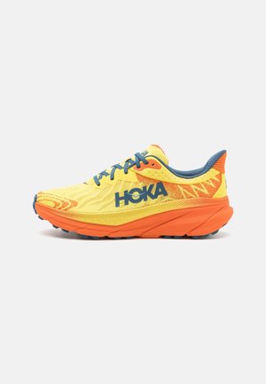 Gele en oranje hardloopschoen met blauwe veters en "HOKA"-logo aan de zijkant, gedempte zool en getextureerde buitenzool voor grip.