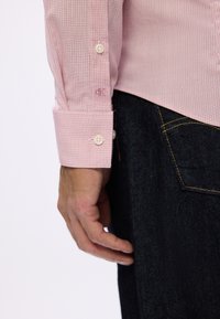 Helles, gestreiftes rosa Hemd mit Knöpfen an den Manschetten, versehen mit einem kleinen "CK"-Logo auf dem Ärmel. Kombiniert mit dunklen Jeans.