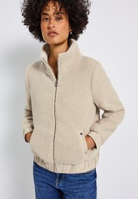 Giacca in pile beige con colletto alto, chiusura a zip, polsi e vita elasticizzati, dotata di tasche laterali e una superficie morbida e testurizzata.