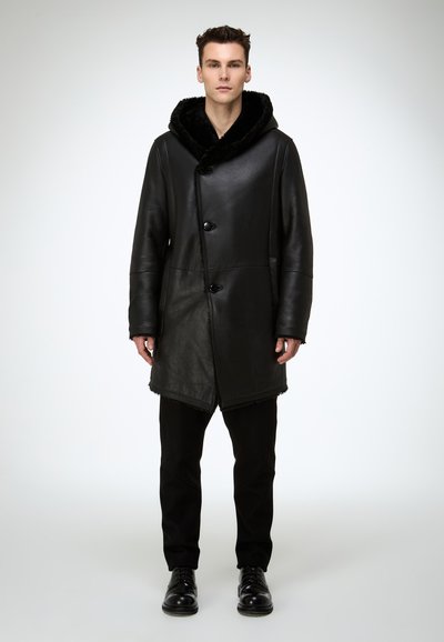 Schwarzer Leder Mantel mit strukturierter, pelzgefütterter Kapuze, Vorderknöpfen und einem leicht asymmetrischen Saum, getragen über schwarzen Hosen und Schuhen.