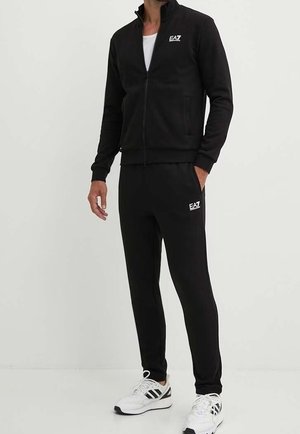Mies, jolla on musta EA7-verryttelypuku vetoketjutakilla ja jogger-housuilla, yhdistettynä valkoisiin Adidas-tennareihin, seisoo yksinkertaisen vaalean taustan edessä.
