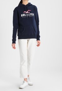 Person som bär en marinblå Hollister-hoodie, vita raka byxor och vita sneakers, står framför en enfärgad ljus bakgrund.