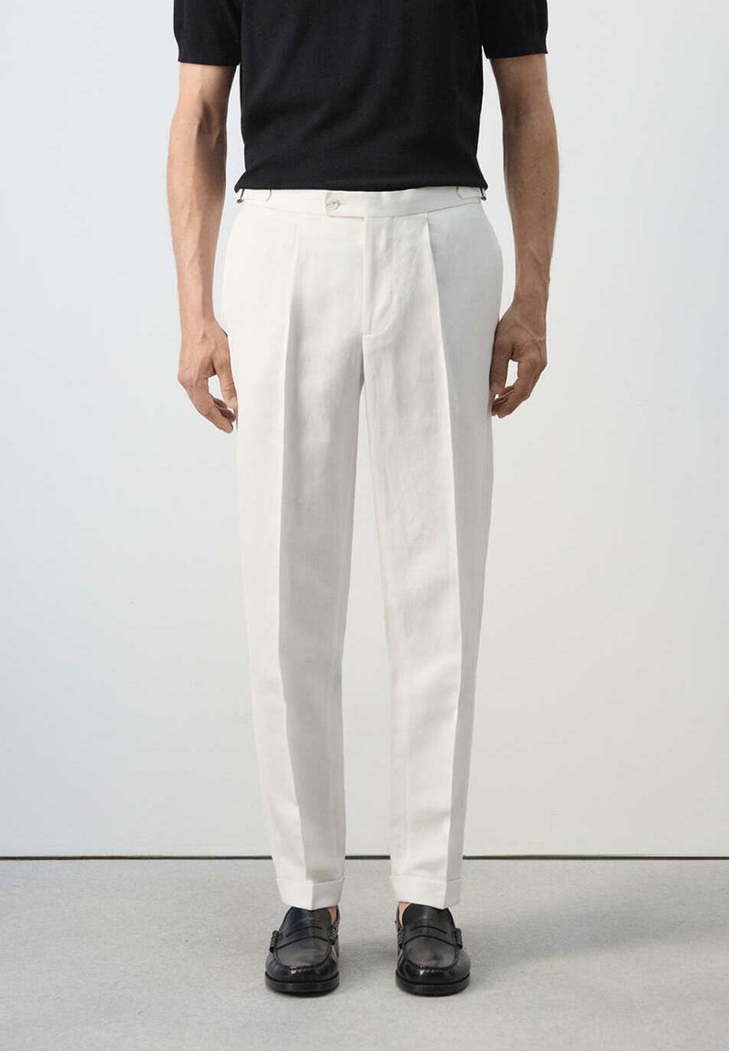 Homme portant un pantalon blanc sur mesure, des mocassins noirs et une chemise noire à manches courtes, debout devant un fond uni.