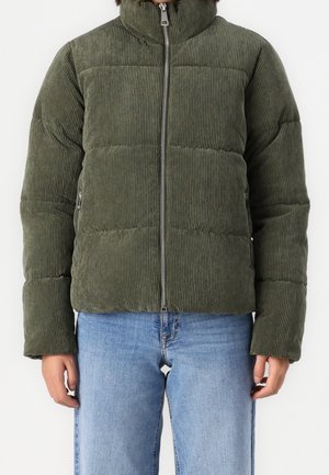 Veste matelassée en velours côtelé vert avec fermeture éclair sur le devant, portée avec un jean bleu clair, visible des épaules aux hanches sur fond blanc.