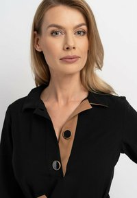 Czarna koszula z kołnierzykiem w kolorze tan z dwoma czarnymi, okrągłymi guzikami. Materiał wydaje się gładki i lekki. Proste elementy wzornicze.