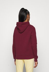 Felpa bordeaux con cappuccio con coulisse, polsini e orlo a costine. Indossata con pantaloni gialli a quadretti, vista da dietro. Tessuto morbido.