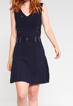 Vrouw draagt een mouwloze marineblauwe jurk met V-hals, decoratieve gespdetails op het lijfje en de taille, staand tegen een effen achtergrond.