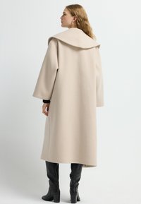 Beige uldblandingsfrakke med en stor, foldet krave og brede ærmer, som går til midt på læggen, parret med knæhøje sorte støvler.