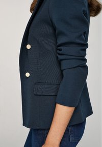 Blazer blu navy in tessuto strutturato, con design doppiopetto, bottoni dorati e una tasca laterale.