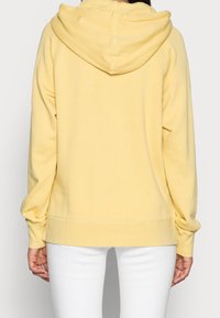 Felpa gialla con tessuto morbido, cappuccio con coulisse e polsini a costine. Abbinata a pantaloni bianchi, vista posteriore.