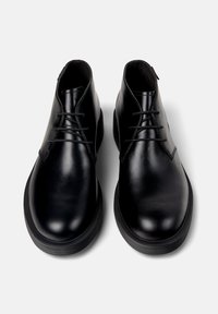 Un par de botas de cuero negro pulido hasta el tobillo con puntas redondeadas y cordones negros, fotografiadas desde arriba sobre un fondo blanco.