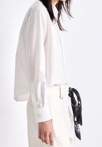 Vit randig skjorta med knappar och långa ärmar, kombinerad med cremefärgade byxor och en svart mönstrad scarf knuten vid midjan.