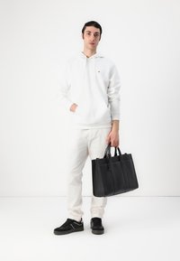 Sac en cuir noir avec des poignées doubles ; texture lisse et design structuré. Porté avec un sweat à capuche blanc et un pantalon clair.