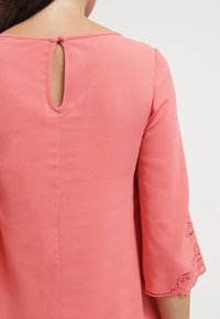 Korálová blůza s třičtvrtinovými rukávy, s výstřihem ve tvaru klíčové dírky na zadní straně a krajkovými detaily na okrajích rukávů. Hladká textura a elegantní design.