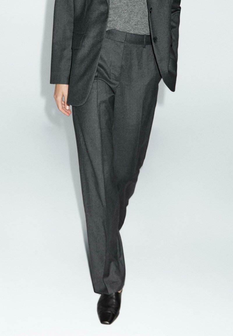 Pantaloni di costume color grigio ardesia realizzati in tessuto pregiato, con vestibilità su misura, pieghe frontali e tasche laterali. Indossati con scarpe eleganti nere.