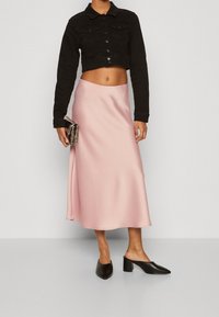 Woman wearing a black cropped jacket, pink satin midi skirt, black heeled mules, holding a metallic clutch purse.