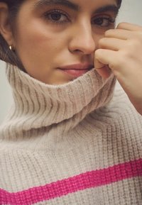 Beige turtleneck sweater med ribbet tekstur og en markant lyserød stribe. Modellens hånd hviler mod kraven, hvilket viser stoffets blødhed.