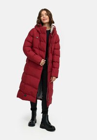 Donna che indossa un lungo cappotto imbottito rosso con cappuccio, pantaloni neri e stivali neri con lacci, in piedi contro uno sfondo neutro.