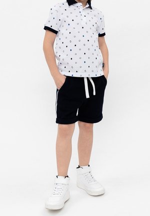 Dziecko ubrane w biały polo w niebieskie i czarne kropki, czarne spodenki ze sznurkiem w pasie oraz białe wysokie trampki, z rękami w kieszeniach.