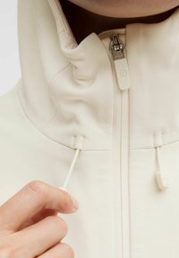 Krémová bunda s vysokým golierom, so zapínaním na zips vpredu a nastaviteľným šnúrkovým pásom. Hladká, mierne textúrovaná tkanina s minimálnymi kovovými doplnkami.