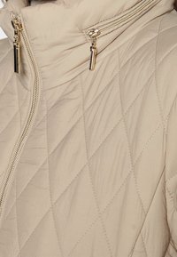 Chaqueta acolchada beige con cuello alto, que cuenta con dos bolsillos con cremallera y detalles en tono dorado. Textura suave con patrón de diamantes.