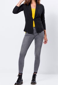 Czarny dopasowany cardigan z głębokim dekoltem w serek i jednym guzikiem, noszony na żółtej bluzce na ramiączkach. Szare obcisłe dżinsy i czarne botki na sznurowadła.