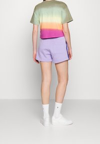 Person som bär en grön till rosa gradient kortärmad t-shirt, lavendelfärgade shorts med bakficka, vita logostrumpor och vita sneakers stående vänd bort.