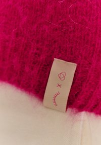 Fuksjowy dzianinowy wełniany materiał o miękkiej fakturze, połączony z kremową bazą tkaniny. Posiada małą beżową metkę z zaokrąglonymi wzorami graficznymi.