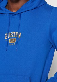 Sudadera azul hecha de una tela suave, con un bolsillo frontal tipo canguro y texto bordado en dorado: "BOSTON USA 1987" encima del bolsillo.