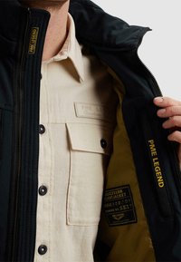 Mann in beige Hemd öffnet schwarze PME Legend Jacke, die gelbes Innenfutter und ein Innenetikett mit Textdetails zeigt.