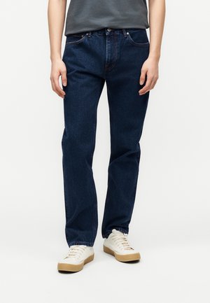 Oseba, oblečena v temno modre kavbojke s širokimi hlačnicami in bele superge, stoji na preprostem svetlem ozadju.