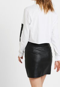 Vit croppad långärmad skjorta med svart grafik på ärmen, tillsammans med en figursydd svart läder minikjol med en slät yta.