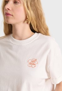 T-shirt in cotone bianco con un ricamo leggero di un fiore di ibisco arancione e la parola "Aloha" sotto. Maniche corte e scollatura rotonda.