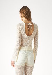 Koronkowy bodysuit w kolorze off-white z długimi rękawami, z odkrytymi plecami zabezpieczonymi trokami, o teksturowanym geometrycznym wzorze, w zestawie z pasującymi szortami.