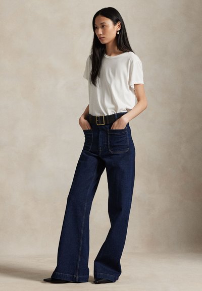 Moteris stovi kišenėse su rankomis, dėvi baltą trumpomis rankovėmis marškinius, tamsius plačius džinsus su priekinėmis kišenėmis, juodą diržą ir juodus batus.